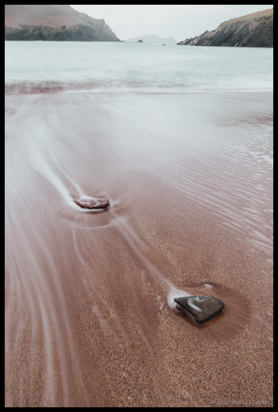 Long exposure photograph of Clogher Strand on the Dingle Peninsula showing silky water flowing around two dark rocks on wet sand, with dramatic coastal headlands and misty sea in the background under an overcast sky.