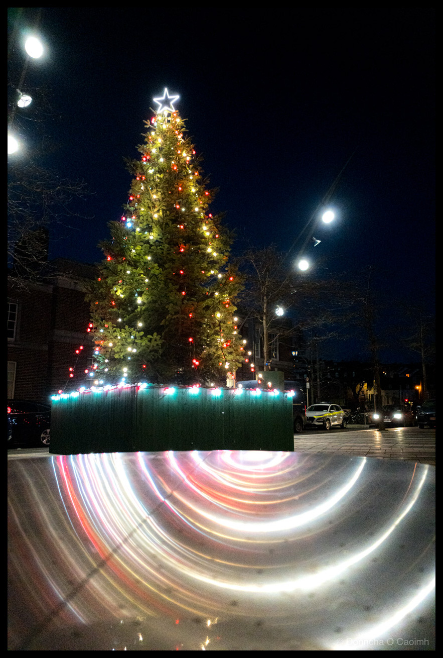 Night photograph of an illuminated Christmas tree on Cornmarket Street, Cork, with multicoloured lights and white star topper, with colourful light trail reflections streaking across a polished steel table surface in the foreground.