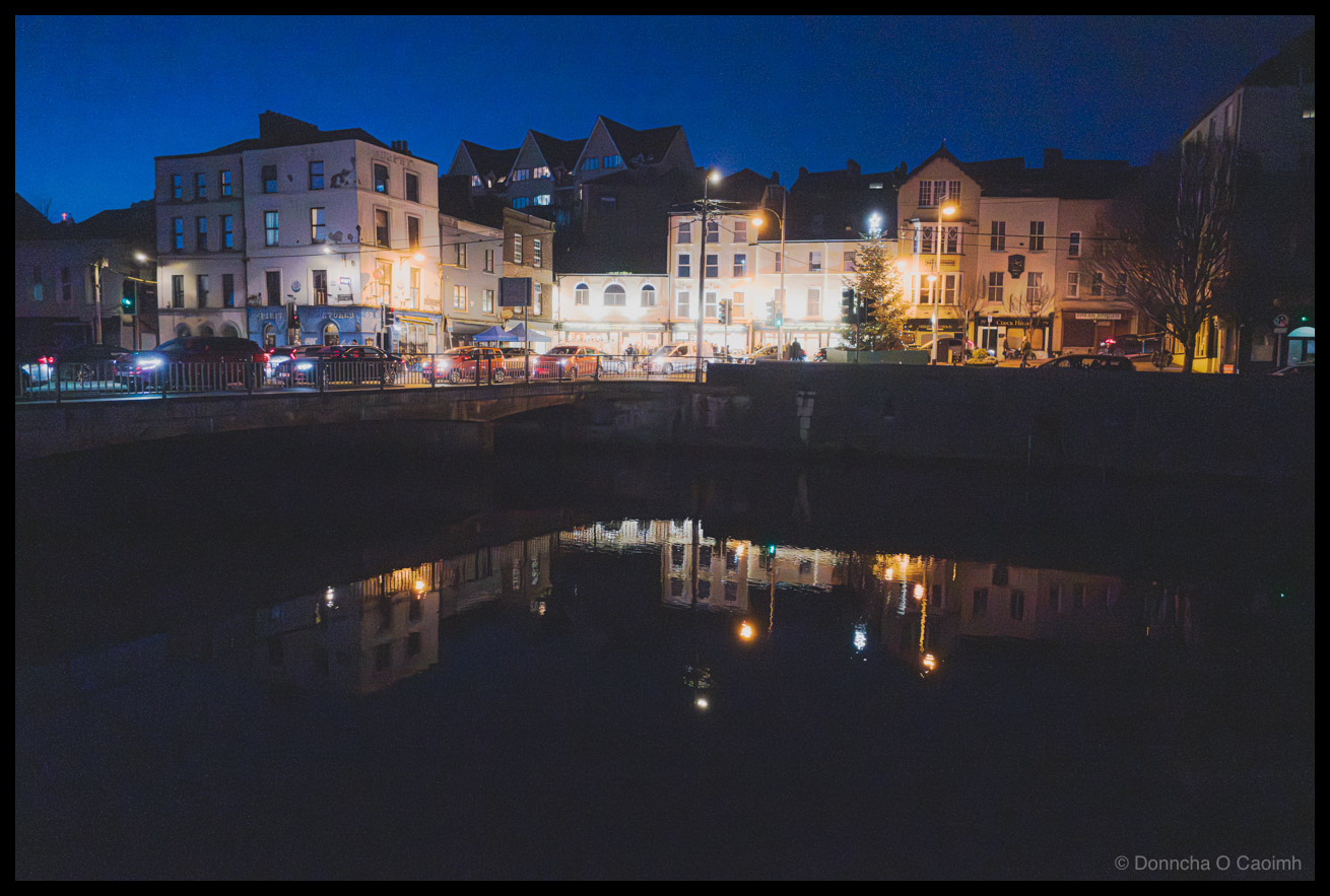 Night photograph of illuminated buildings along Farren's Quay at the end of Shandon Street in Cork, with their warm lights and a small Christmas tree reflecting in the still dark waters of the River Lee during blue hour.
