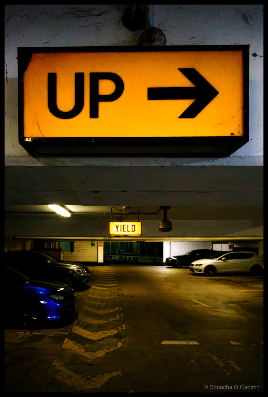 Interior of North Main Street car park in Cork showing an illuminated orange "UP" directional sign with arrow above a yellow "YIELD" sign, with parked cars and weathered concrete ceiling visible in the ambient lighting.