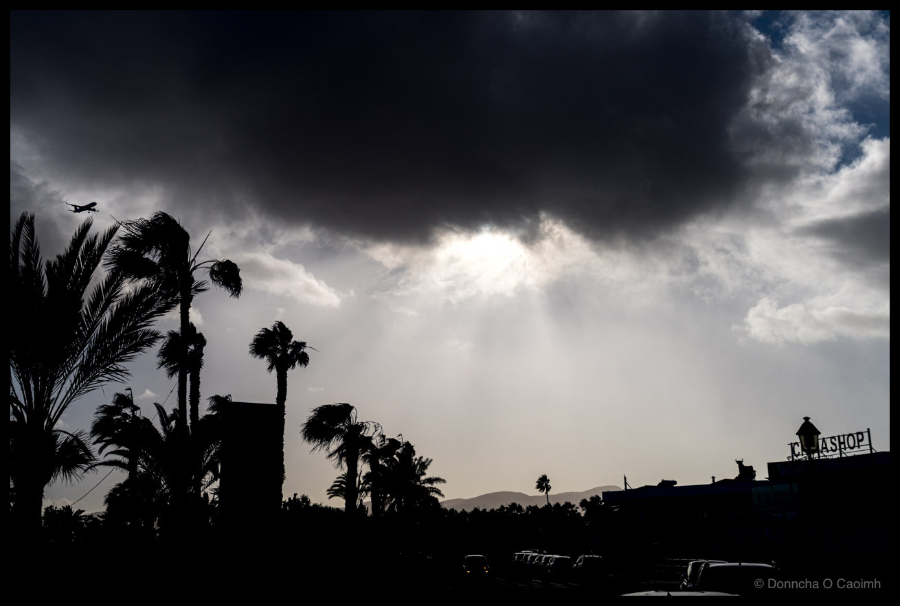 Dramatic black and white photograph of a plane descending against dark storm clouds over Castillo Caleta de Fuste, Fuerteventura, with palm trees silhouetted in the foreground and a shop sign visible on the right.