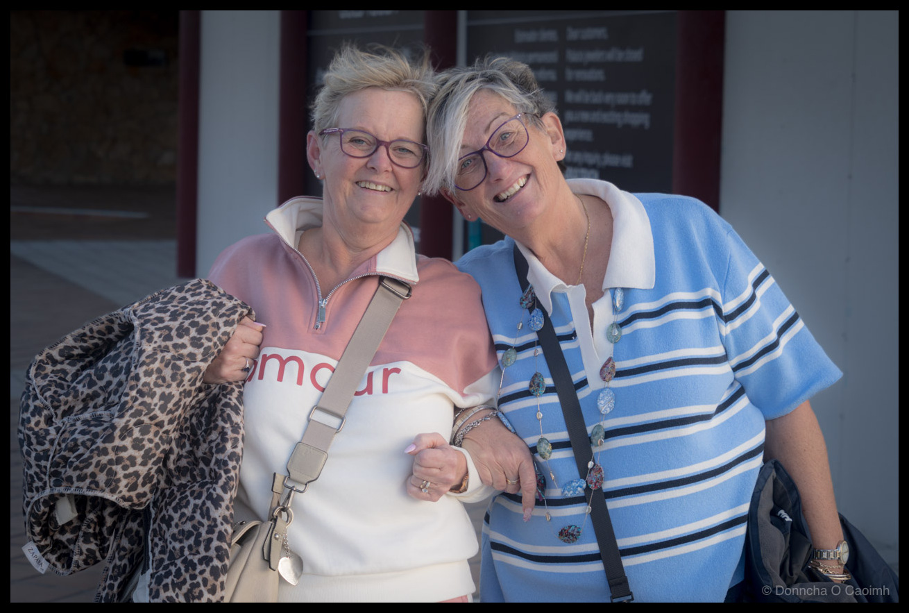 Two cheerful women with grey hair and glasses posing together with arms around each other in Castillo Caleta de Fuste, Fuerteventura, one wearing a pink hoodie with "amour" text and leopard print jacket, the other in a blue and white striped shirt, both smiling broadly at the camera.