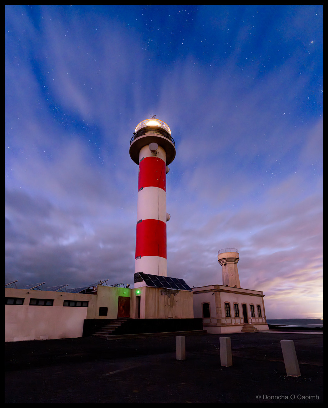 The illuminated Faro de Tostón lighthouse with distinctive red and white horizontal stripes against a deep blue twilight sky filled with stars during blue hour in Fuerteventura, with white buildings and solar panels at the base and pink gradient clouds near the horizon.