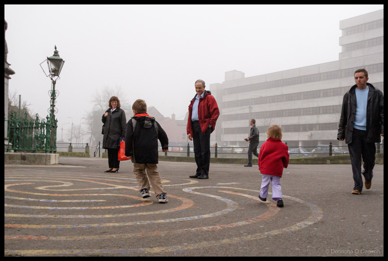 Children playing on a circular painted maze on the pavement near Cork's National Monument on a foggy day, with adults watching and the old tax office visible in the misty background, with an ornate Victorian lamp post on the left.