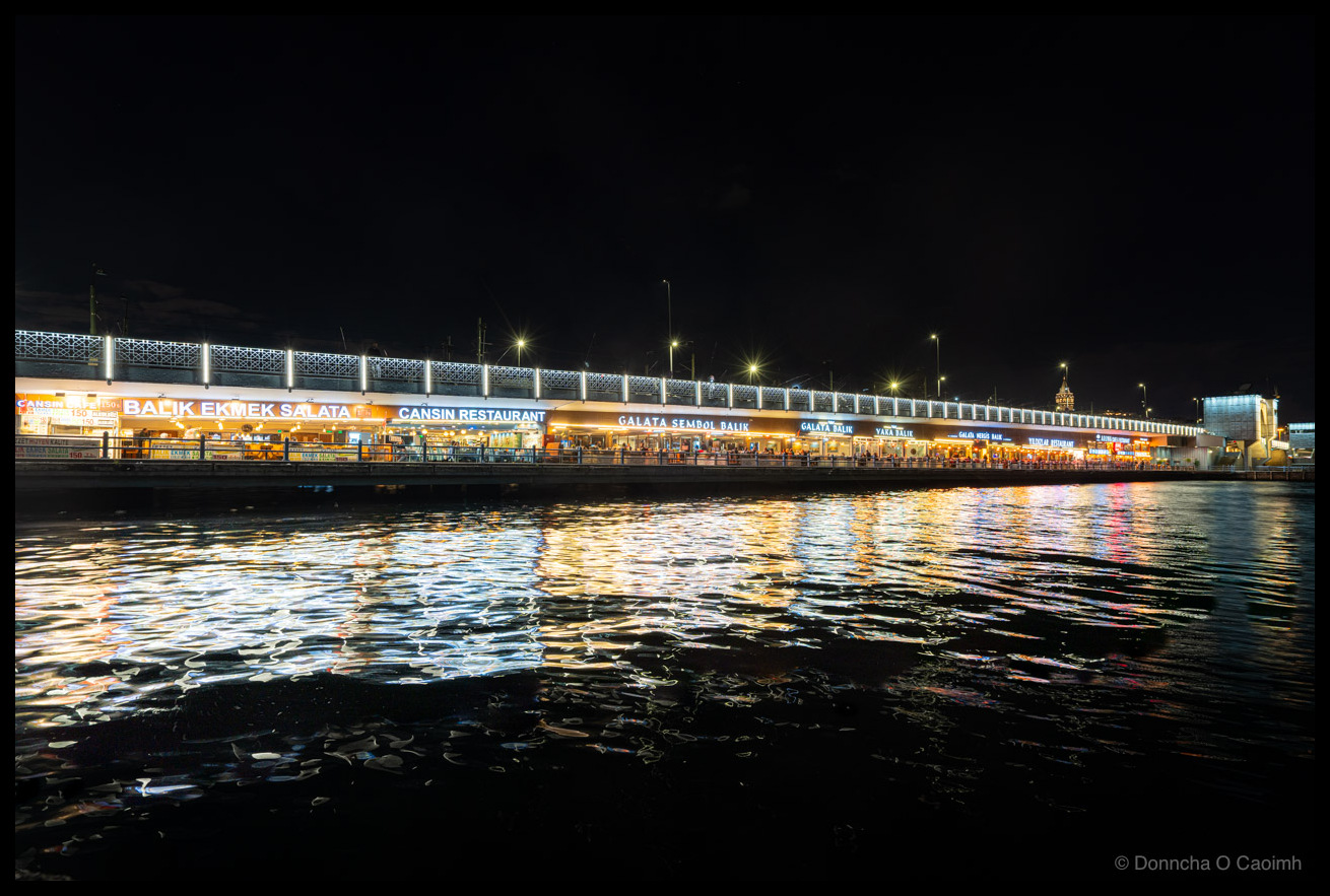 Night HDR photograph of Istanbul's illuminated Galata Bridge spanning the Golden Horn, with Turkish fish restaurants and shops on the lower level reflecting in the rippling water below, street lights creating golden trails, and historic buildings visible in the background.