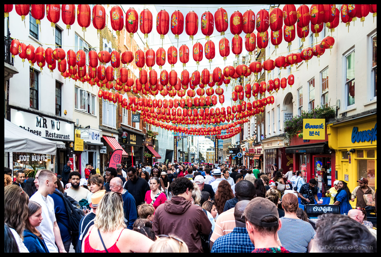 Photograph of a densely packed crowd of people walking through London's Chinatown on Wardour Street, with dozens of red and gold Chinese lanterns strung overhead in multiple rows between the buildings. Visible shopfronts include MR WU Chinese Restaurant with a HOT BUFFET sign on the left, and the yellow-fronted Bunsik (분식) Korean restaurant on the right. A Rupert Court street sign is visible on the right-hand building. The Chinatown gate is just visible in the far distance. An LNER advertising banner hangs among the lanterns. O'Neills pub can be seen at street level on the right.