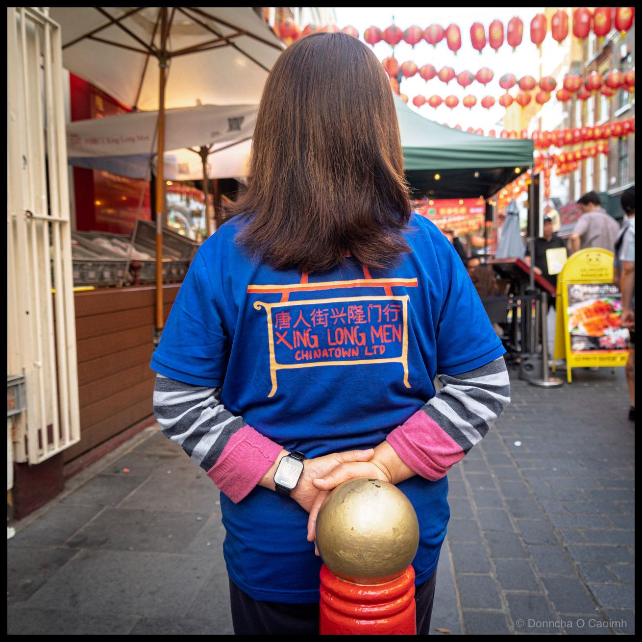 Photograph taken from behind of a woman standing in London's Chinatown on a July evening, wearing a blue t-shirt over a grey and pink striped long-sleeve top. The back of the t-shirt reads 唐人街兴隆门行 XING LONG MEN CHINATOWN LTD with an illustration of the Chinatown gate. Her hands are clasped behind her back, resting on a red bollard with a gold ball on top. She wears a smartwatch on her left wrist. Red lanterns are strung overhead, and restaurant parasols, food stalls, menu boards, and other pedestrians are visible in the background.