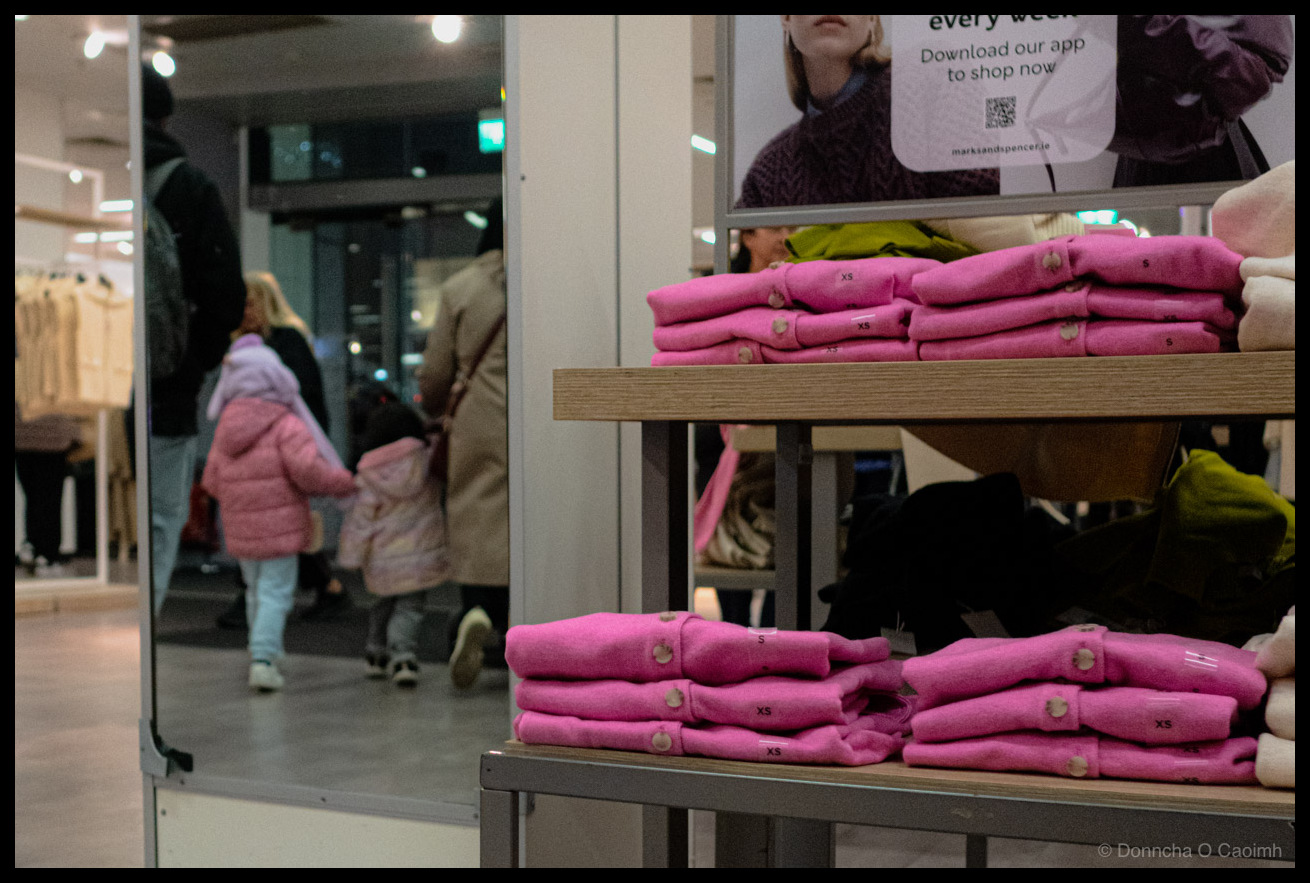 A child in a pink puffer jacket walking out of a clothing shop with an adult, seen in a mirror next to display shelves stacked with bright pink folded jumpers in matching shades, with shop interior and mirrors visible in the background.