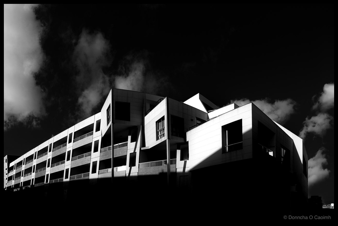 Dramatic high-contrast black and white panoramic photograph of the Eurostars Las Salinas hotel in Castillo Caleta de Fuste, Fuerteventura, with stark white modern architecture featuring angular balconies against an ominous dark sky with billowing clouds.