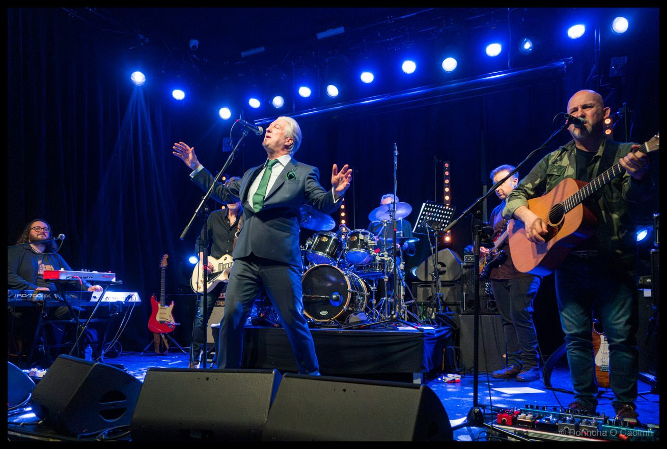 A David Bowie tribute band performing on stage at Cyprus Avenue in Cork under blue stage lights, with the lead singer, Peter Quinn, in a dark suit and white hair singing with expressive hand gestures, flanked by band members on keyboards, drums, and acoustic guitar.