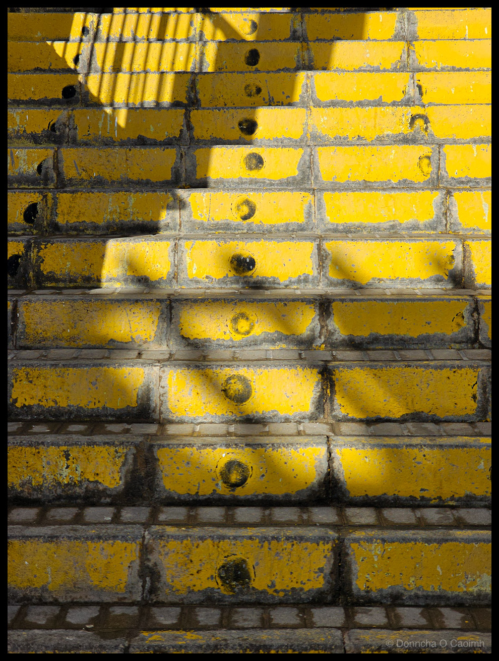 Close-up photograph of steep yellow-painted concrete steps with dramatic diagonal shadows, weathered paint, dark stains, and a corrugated pipe visible at the top, leading to a holiday apartment in Fuerteventura.