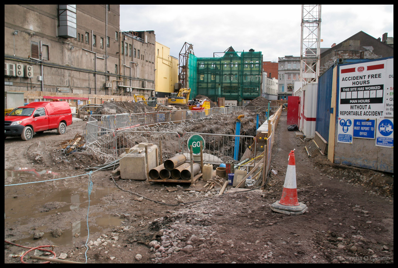 Photograph of the muddy construction site of the Dunnes Stores development on St Patrick's Street, Cork, taken on 17 March 2008. The site is a landscape of churned earth, rubble, metal barriers, concrete pipes, puddles, and a traffic cone. A red Toyota Hilux pickup is parked to the left. Yellow excavators are visible in the middle distance, and the green-netted steel frame of the new building rises in the centre background. A Sisk construction company sign on the right reads ACCIDENT FREE HOURS THIS SITE HAS WORKED MAN HOURS WITHOUT A REPORTABLE ACCIDENT, alongside a blue ALL VISITORS MUST REPORT TO OFFICE sign and mandatory safety equipment notices. Site cabins and older commercial buildings line the edges of the site under an overcast sky.