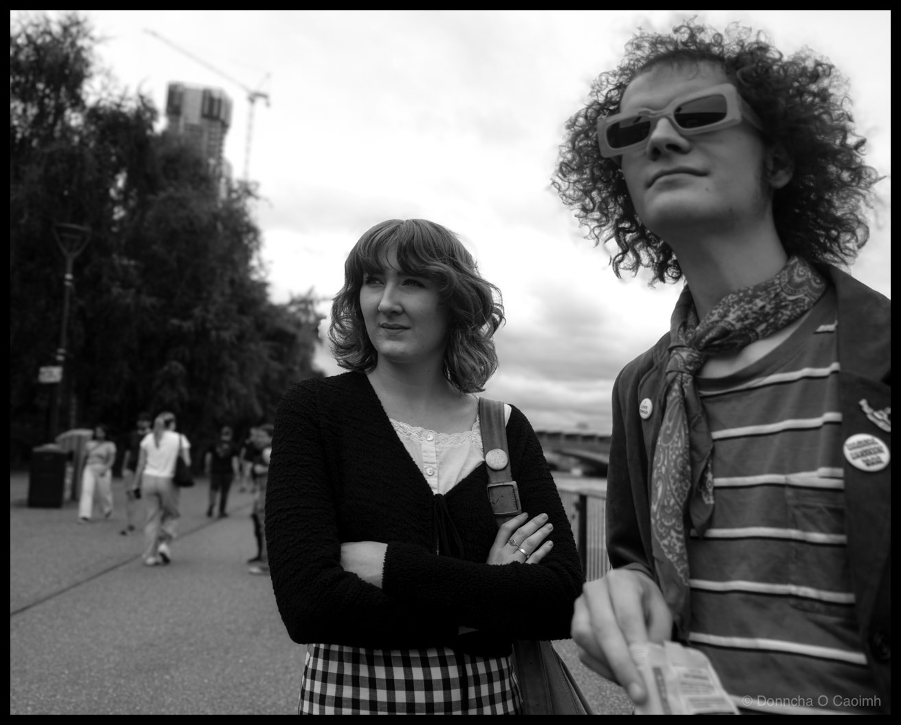 Black and white photograph of two young people standing on the South Bank near the Millennium Bridge in London. A woman with wavy shoulder-length hair stands with arms folded, wearing a black cardigan over a white lace-trimmed top and a black and white gingham skirt, with a small bag over her shoulder. To the right, a young man with a large curly afro wears sunglasses, a paisley scarf, and a striped shirt with small pin badges, holding what appears to a leaflet. Other pedestrians, trees, street lamps, and buildings are visible in the soft-focus background.