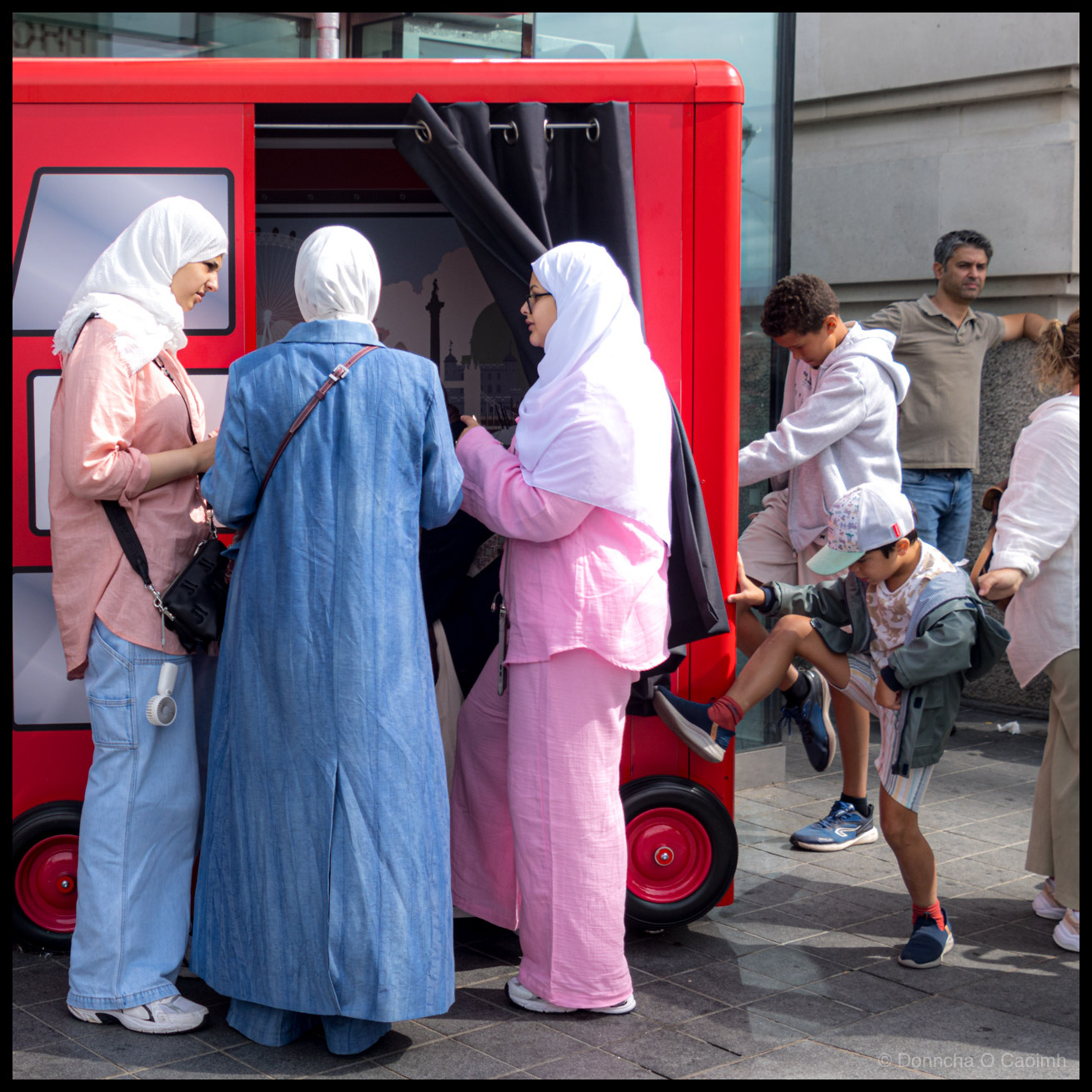 Photograph of people gathered around a red novelty photo booth shaped like a small vehicle on a London street. Three women wearing white headscarves stand at the booth's curtained entrance, one in a pink top and jeans, one in a long blue dress, and one in a pink outfit. A boy in a grey hoodie and a younger boy in a green cap and grey jacket plays with the red wheels on the side. Other pedestrians stand nearby. A London landmark backdrop is visible through the booth's window.
