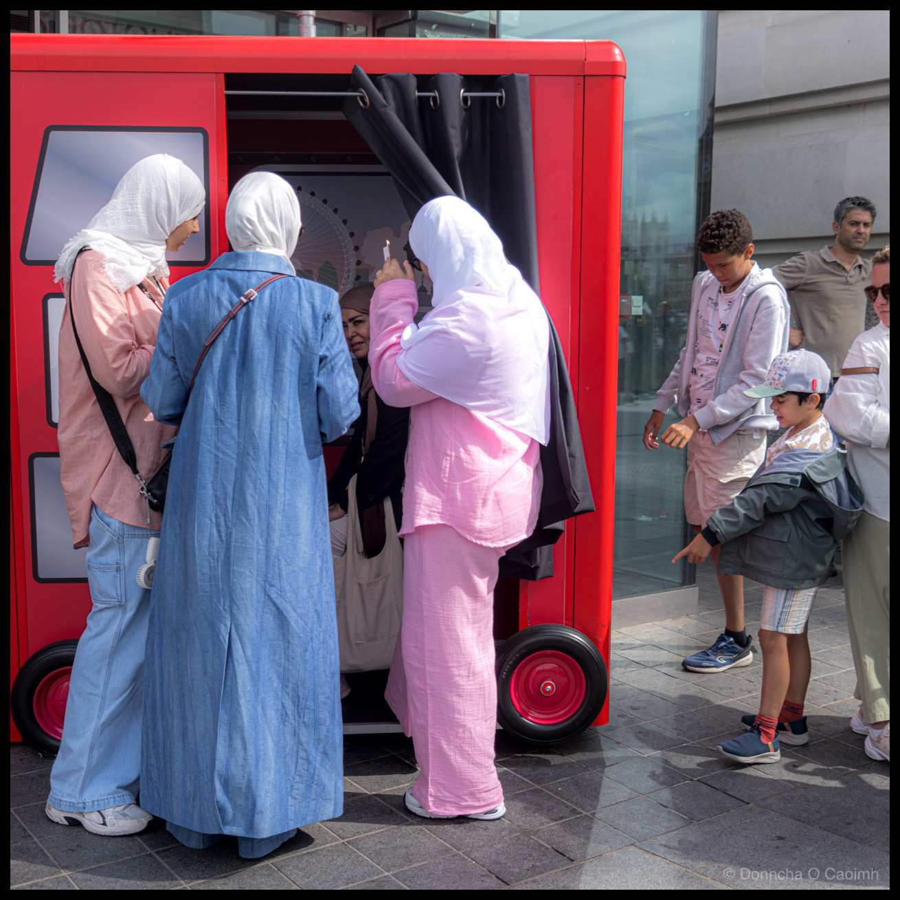 People at a red novelty photo booth. The three women in headscarves are gathered at the entrance, with one holding up her phone to photograph whoever is inside the booth. A fourth woman is now visible seated inside. The younger boy in the green cap is laughing and pointing at the wheels of the booth while the older boy in the grey hoodie watches. A man in a beige polo shirt stands to the right looking on.