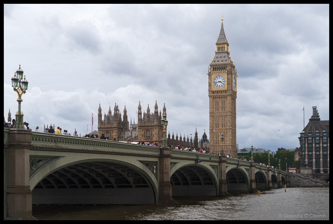 Photograph of Westminster Bridge spanning the River Thames with the Elizabeth Tower (Big Ben) and the Palace of Westminster behind it under a dramatic cloudy sky. Pedestrians crowd the bridge, and an ornate green and gold Victorian lamp post stands on the near side. The Gothic pinnacles and towers of the Houses of Parliament stretch across the background, with a red bus just visible on the far side of the bridge. The murky green-brown water of the Thames fills the foreground.