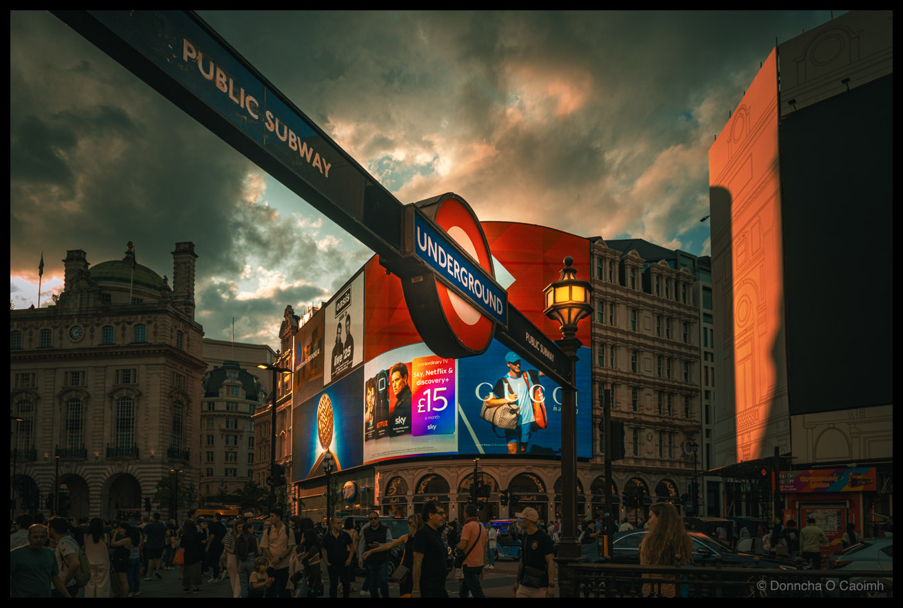 Photograph of Piccadilly Circus in London at dusk, with the iconic London Underground roundel sign and a PUBLIC SUBWAY sign in the foreground. The large curved LED advertising screens display adverts for Sky, Netflix & discovery+ at £15 a month, Gucci, and live25. A warm glowing street lamp sits beside the Underground sign. Dramatic storm clouds are lit from below by the orange glow of the setting sun and the LED screens. Crowds of pedestrians fill the pavement below, with the ornate Victorian and Edwardian buildings of Piccadilly Circus surrounding the scene.
