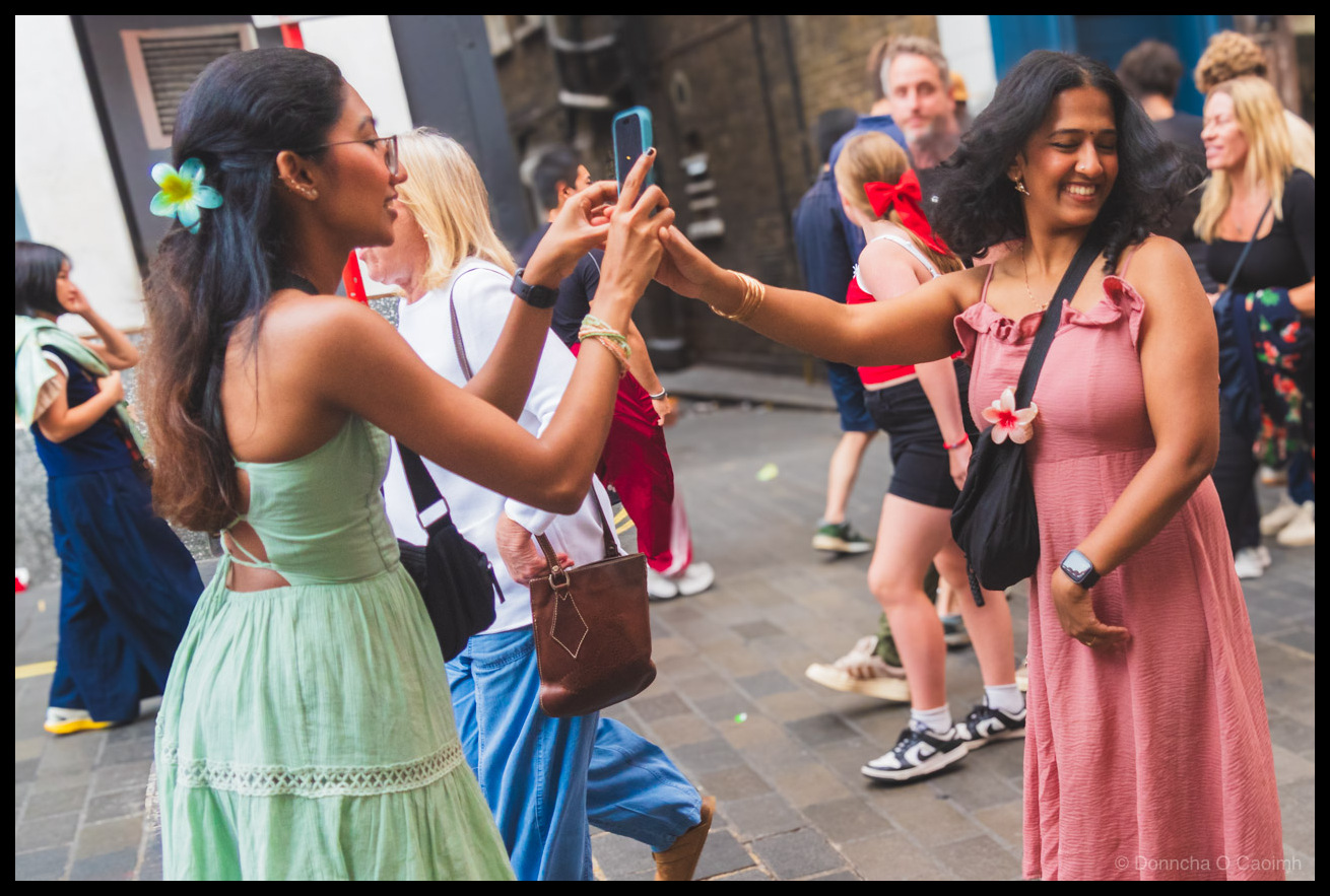 Photo of a woman in a green tiered dress with a flower in her hair, holding up a phone in a turquoise case to photograph another woman in a pink ruffled dress who is smiling back at her. Both wear bracelets and the woman in pink has a smartwatch and a flower pinned to her black bag strap. A busy London street scene fills the background with other pedestrians.