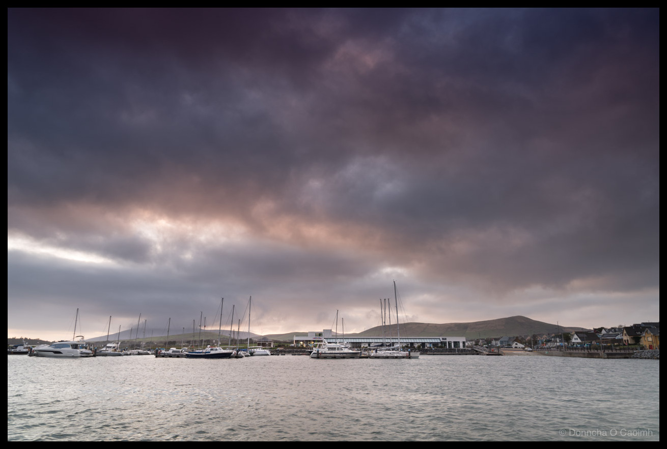 Photo of Dingle Marina viewed from the pier in Dingle, with sailboats and motor cruisers moored along the pontoons beneath a dramatic sky of dark purple and grey storm clouds with hints of pink. Green hills rise behind the marina buildings and houses line the waterfront to the right.