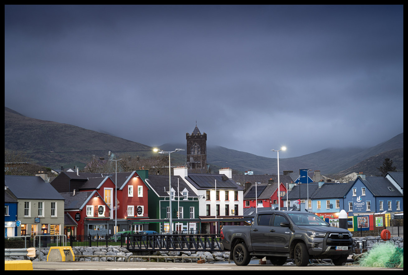 Photo of Dingle town at dusk, with a row of colourful shopfronts in red, green, white, and blue beneath a heavy grey sky and cloud-covered mountains. A stone church tower rises above the rooftops in the centre. A dark grey Toyota Hilux pickup truck is parked in the foreground near the harbour wall, with metal railings, rocks, and green fishing net visible. Street lights are lit.