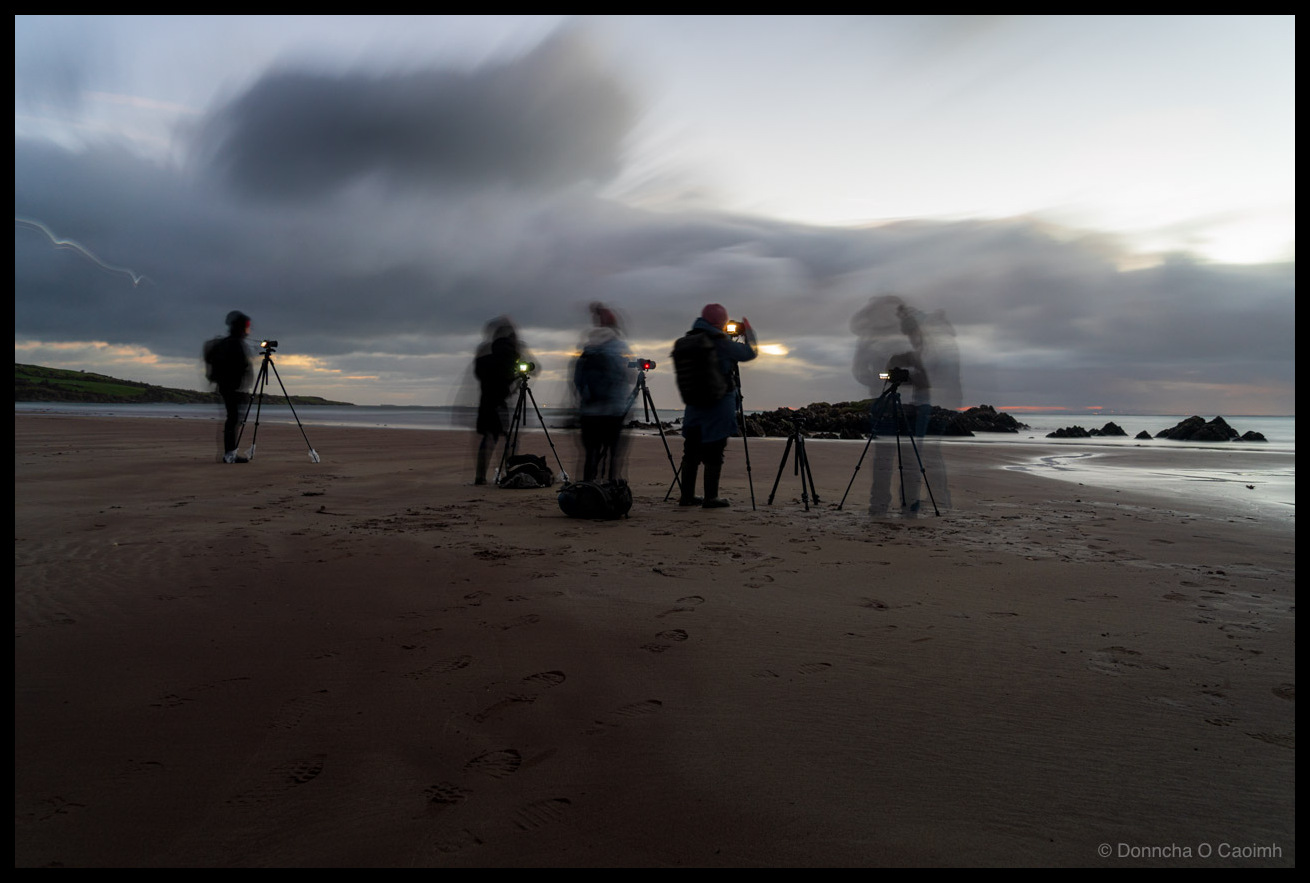 Long exposure photo of a group of photographers with tripods on a dark sandy beach at dawn, their figures rendered as ghostly blurs by the slow shutter speed. Cameras on tripods remain sharp while the people appear translucent against a dramatic sky of streaked clouds. A streak of light on the left is another photographer with a light on their head walking away. Rocks jut from the shoreline to the right.