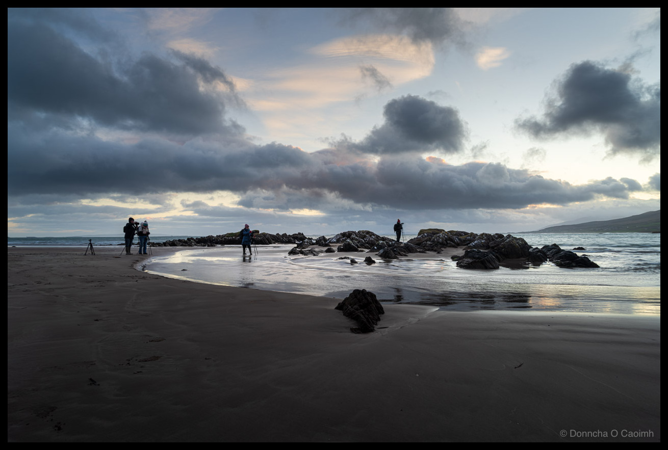 Photo of photographers with tripods spread across a beach at dawn, some clustered near jagged dark rocks while one stands alone on a rocky outcrop to the right. Wet sand reflects a sky of layered grey and pink clouds. Mountains are visible on the far right horizon. Footprints mark the sand in the foreground.