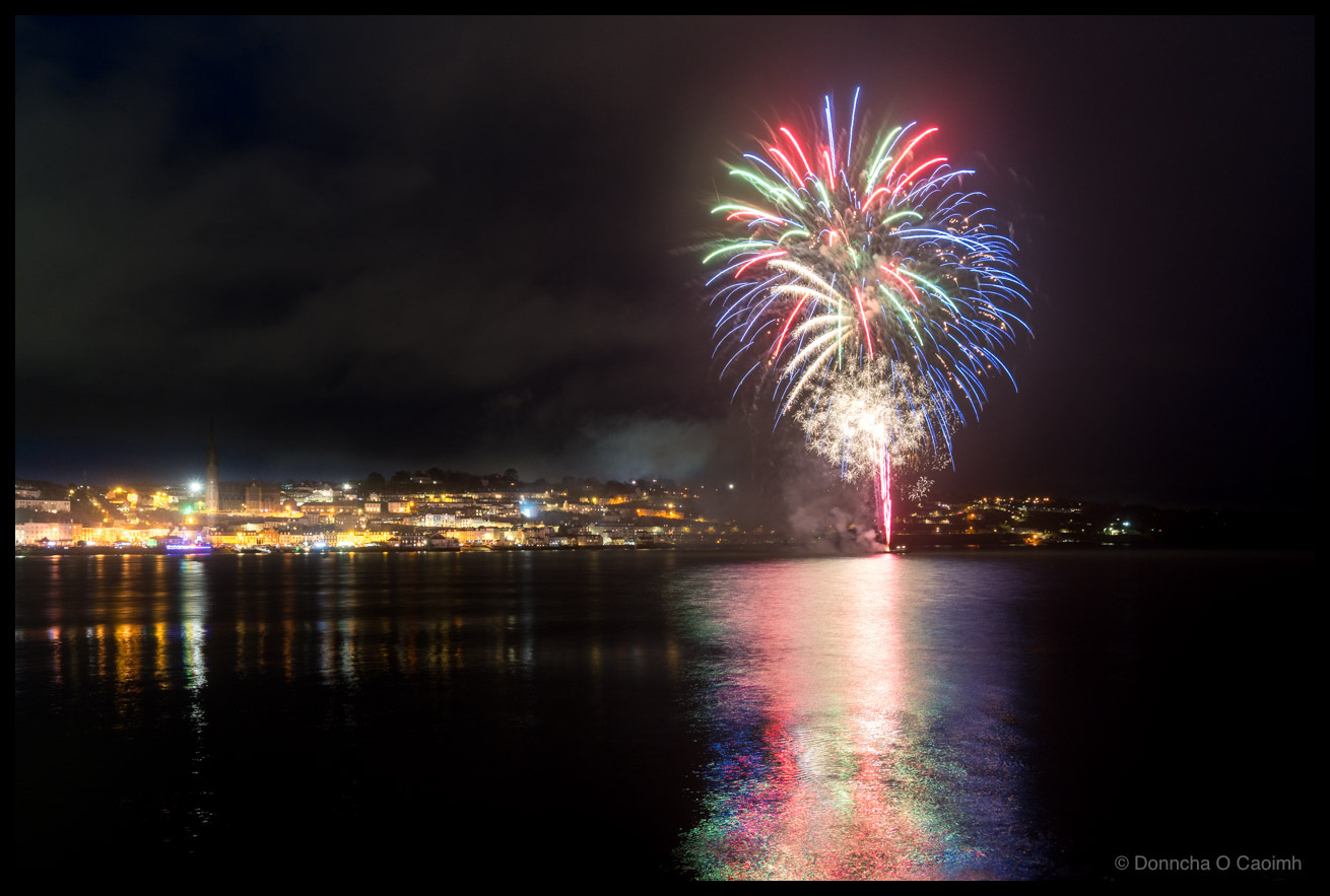 Photo of a spectacular fireworks display over Cobh harbour at night, with bursts of green, red, blue, and white light cascading above the town's illuminated waterfront. The fireworks and town lights reflect vividly in the still water of Cork Harbour. The spire of St Colman's Cathedral is visible among the lit buildings on the hillside.