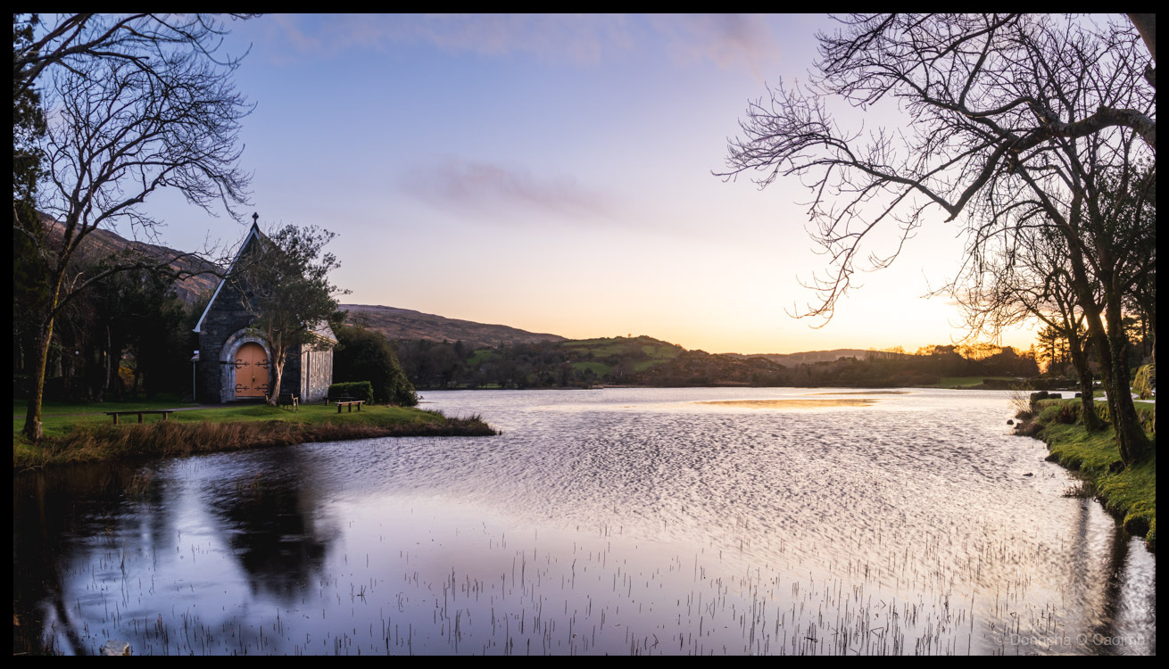 Photo of St. Finbarr's Oratory at Gougane Barra, County Cork, at sunrise, with the stone chapel reflected in the still lake, reeds poking through the water, bare winter trees framing the scene, and hills rising behind under a soft pink and blue sky.