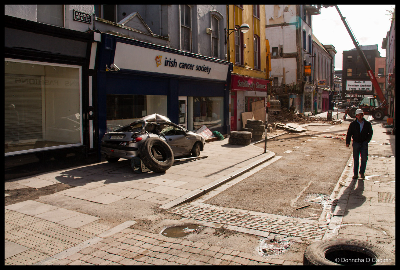 Castle Street in Cork with a black-and-white "CASTLE STREET" street sign on the wall, showing the "irish cancer society" shopfront, "Satellite Dry Cleaners", a "Suits & Leather Jackets From €49.95" sign further down, the crushed silver Peugeot with a tyre on its roof on the pavement, a man in a white hard hat and dark jacket walking towards the camera, rubble across the road, and a second tyre in the foreground.