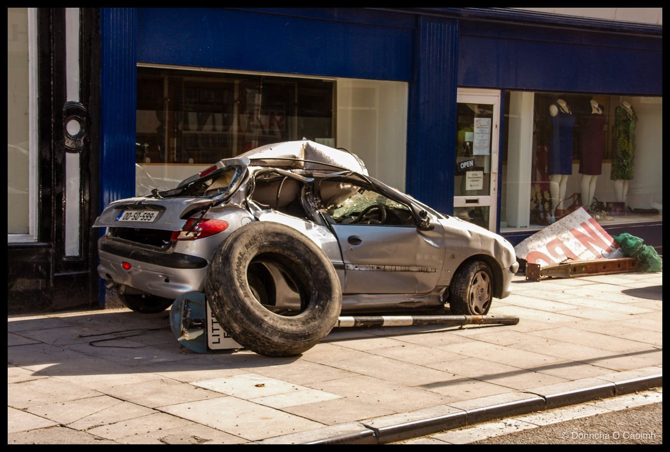A silver Peugeot 206 with Irish registration "00-SO-599" crushed flat from above, its roof caved in and a large lorry tyre resting on the wreckage, parked on a pavement in front of a blue-fronted shop displaying mannequins in dresses with an "OPEN" sign on the door and a toppled red-and-white "INFO" sign lying beside it.