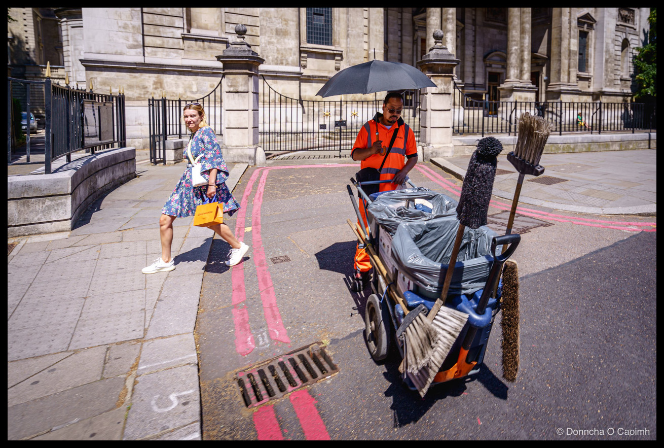 Photo taken on a bright summer day outside the Brompton Oratory in London, showing a street sweeper in a high-visibility orange vest and sunglasses standing beside a cart loaded with bin liners and a row of upright brooms, holding a large black umbrella for shade, while a woman in a blue floral dress, white trainers, a white shoulder bag and holding a yellow bag walks past on the pavement and glances back over her shoulder, with the classical Portland stone façade and iron railings of the church behind them and bright pink survey-style lines painted across the road.