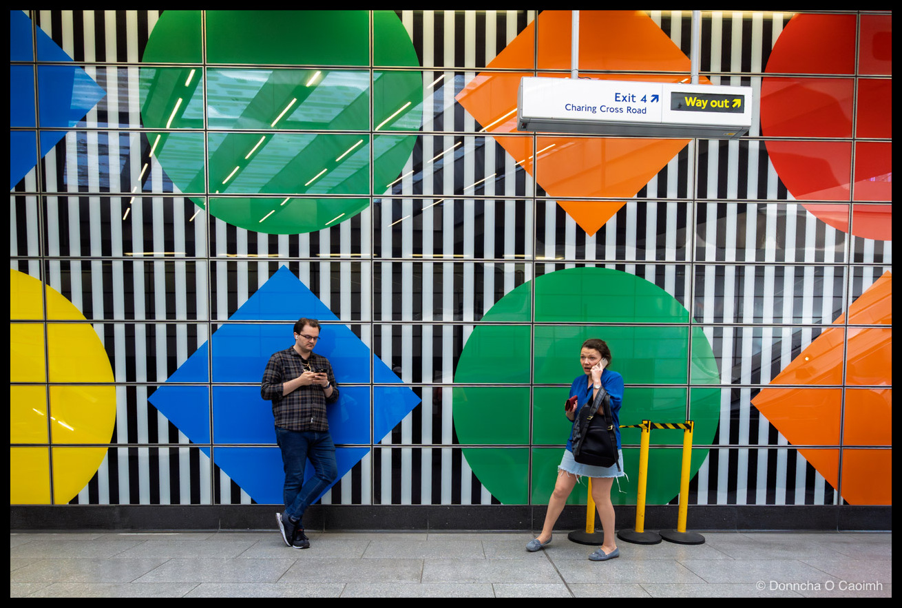 Two people on their phones at Tottenham Court Road Underground station, standing in front of Daniel Buren's "Diamonds and Circles" artwork. It's a wall of large coloured circles and diamonds (yellow, green, blue, orange, red) overlaid on bold black-and-white vertical stripes. A suspended sign above reads "Exit 4 Charing Cross Road" and "Way out". A man in a checked shirt and jeans leans against the wall reading his phone; a woman in a blue jacket and denim skirt holds a phone to her ear, with yellow stanchions and a black bag at her side. Tiled floor in the foreground.