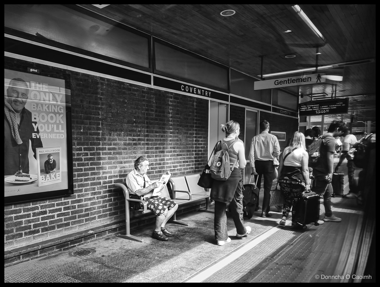 Black and white photo of a platform area at Coventry railway station, with an older man in a shirt, tie and checked shorts sitting on a bench reading a newspaper, a large advertising poster on the wall to his left showing Paul Hollywood and the text "THE ONLY BAKING BOOK YOU'LL EVER NEED" alongside a smaller image of the book "BAKE" by Paul Hollywood, a "COVENTRY" sign on the wall above the bench, a "Gentlemen" sign for the toilets on the right, a digital departure board, and a queue of travellers with suitcases walking towards the platform.