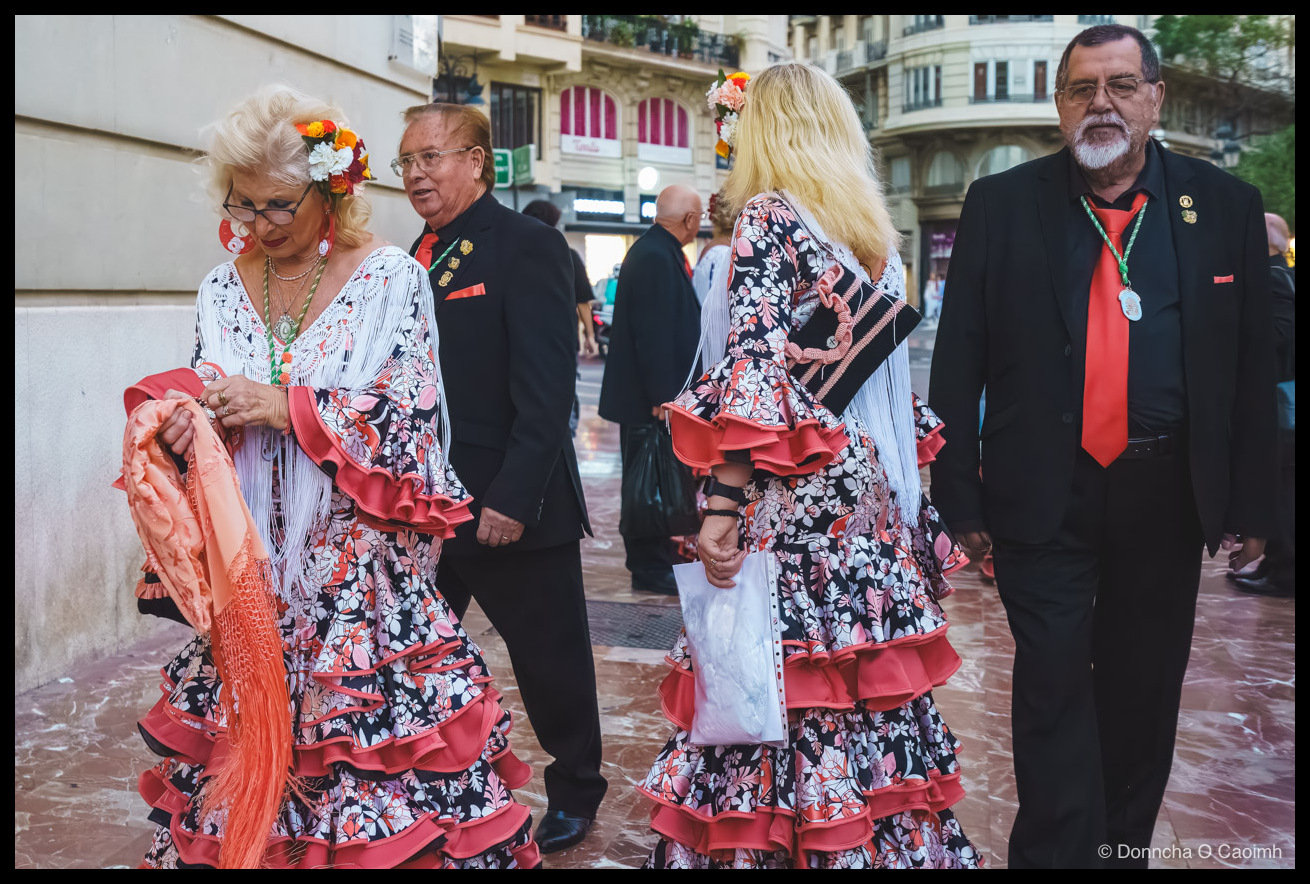 Two women wearing flamenco dresses with floral headpieces walking alongside two men in dark suits with red ties and red pocket squares on a wet, polished street in Valencia, with classical Spanish architecture in the background