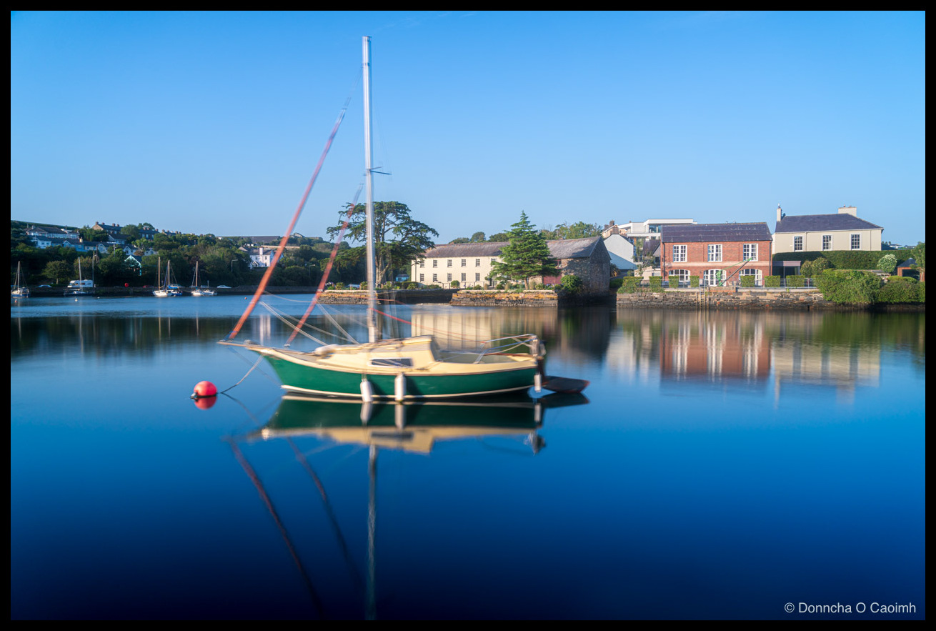 A small green and yellow sailboat moored in the calm waters of Kinsale harbour, with a red mooring buoy beside it, mirror-like reflections in the water, and a row of houses, trees, and other moored yachts along the far shore under a clear blue sky.