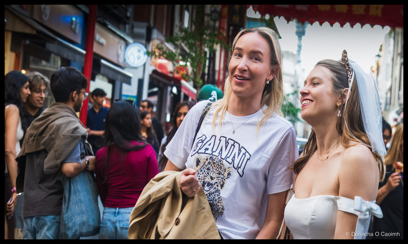 Two women in the foreground on a busy London Chinatown street; the woman on the left has long blonde hair and wears a white t-shirt printed with "GANNI" above a leopard-cub graphic, with a tan jacket draped over her arm; the woman on the right wears a white off-shoulder dress with a small white bow on the sleeve, a leopard-print headband and a short bridal veil; behind them a crowd of pedestrians fills a sunlit street, with red Chinese signboards and lanterns hanging above.