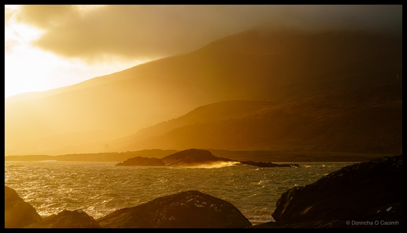 A golden sunrise at Cappagh Beach, County Kerry, with warm light spilling across choppy Atlantic waters and silhouetting dark rocky outcrops in the foreground, while layered mountain ridges fade into haze and mist under dramatic clouds.