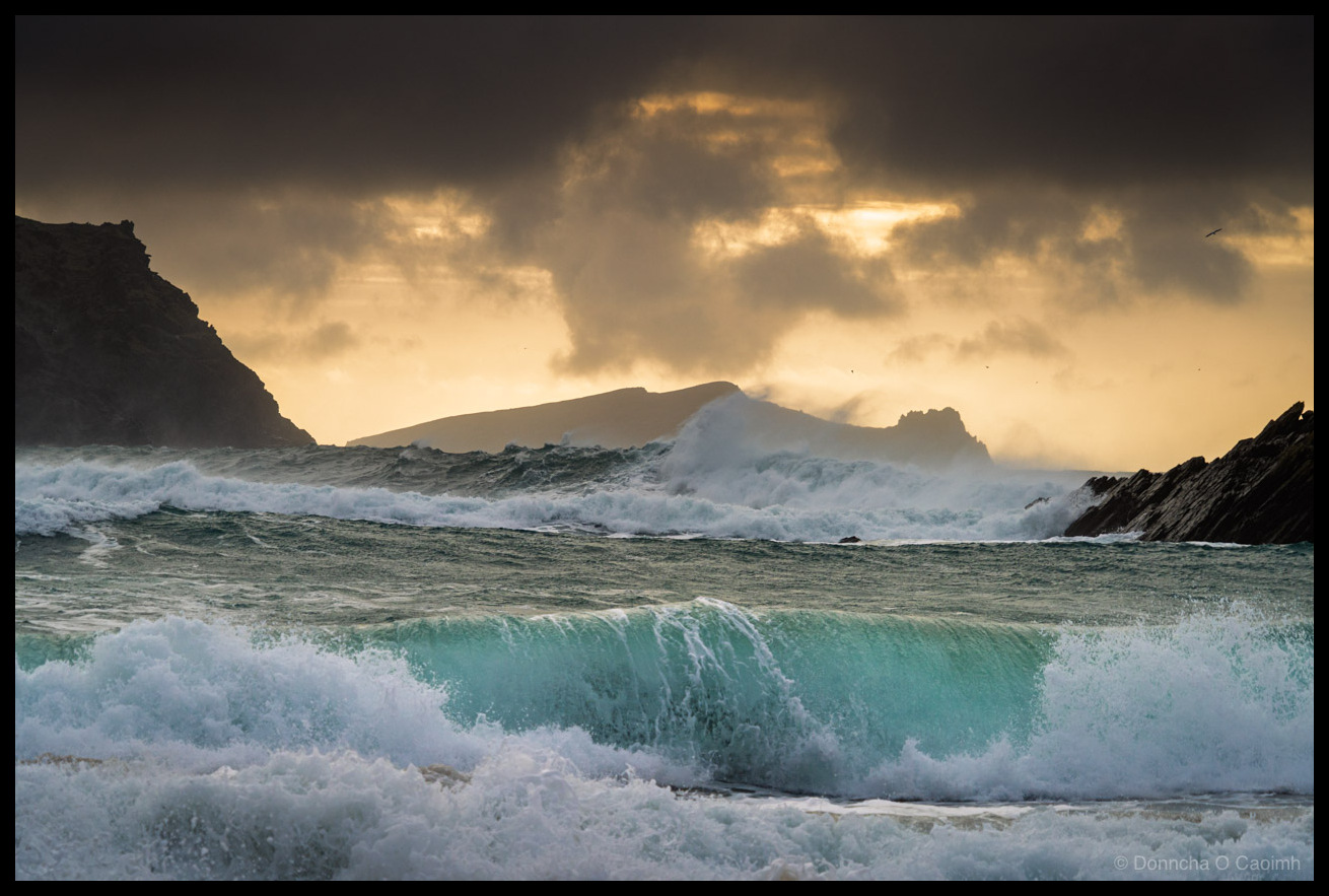 Photo of powerful turquoise waves crashing at Clogher Beach, County Kerry, with the silhouette of An Fear Marbh island visible through sea spray against a dramatic sky of dark storm clouds broken by warm golden light. Dark rocky headlands frame both sides of the scene.