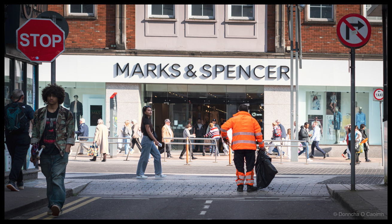 A street cleaner in bright orange hi-vis jacket and trousers bending to pick up a black bin bag on a pedestrian crossing, with shoppers walking past a Marks & Spencer storefront on a busy Cork street; visible text includes a red STOP sign, a no-right-turn sign, "MARKS & SPENCER", "bento", "M&S", "TAXI".
