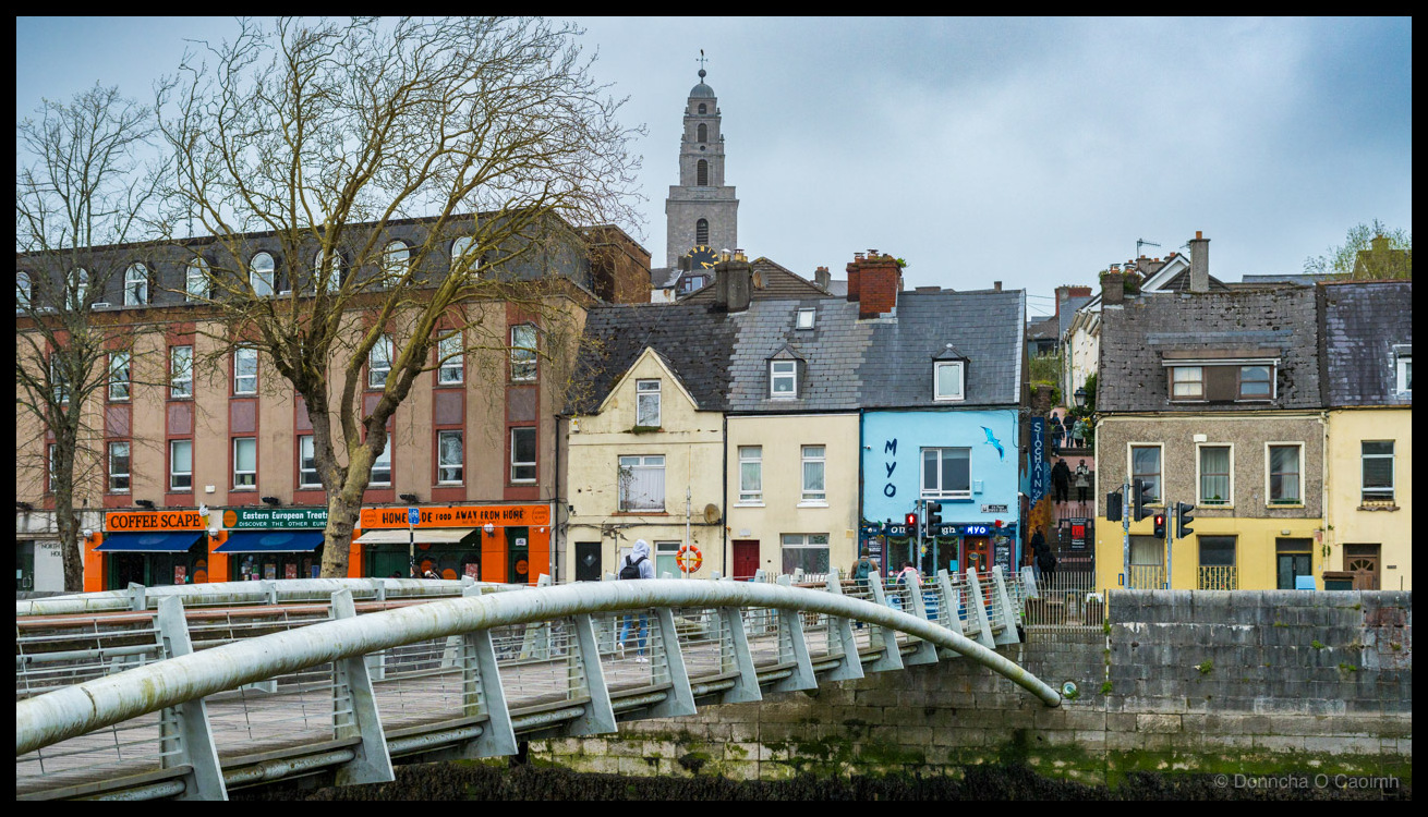 Photo of Cork city's Pope's Quay on an overcast day, with a modern curved pedestrian footbridge in the foreground crossing the River Lee, and a row of colourful buildings behind it including orange-fronted shops reading "COFFEE SCAPE", "Eastern European Treats", "DISCOVER THE OTHER EURO", "HOME MADE FOOD AWAY FROM HOME", a blue-painted building with "MYO" and a painted bird, and a yellow building partially reading "SLOANS".
The tower of St Anne's Church, Shandon rises above the rooftops. A life ring hangs on the quay wall and a figure in a hooded jacket crosses the bridge.