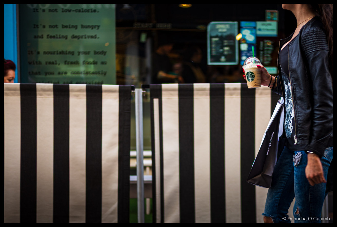 A woman in a black leather jacket and ripped blue jeans walking past Rocket Man, a salad and juice bar in Cork, holding a clear plastic Starbucks Frappuccino cup with the green twin-tailed siren logo and carrying a black newlook.com shopping bag, beside a black-and-white striped barrier; the café window behind her shows menu boards in reflection and a wellness slogan reading "It's not low-calorie. It's not being hungry and feeling deprived. It's nourishing your body with real, fresh foods so that you are consistently satisfied and energised".