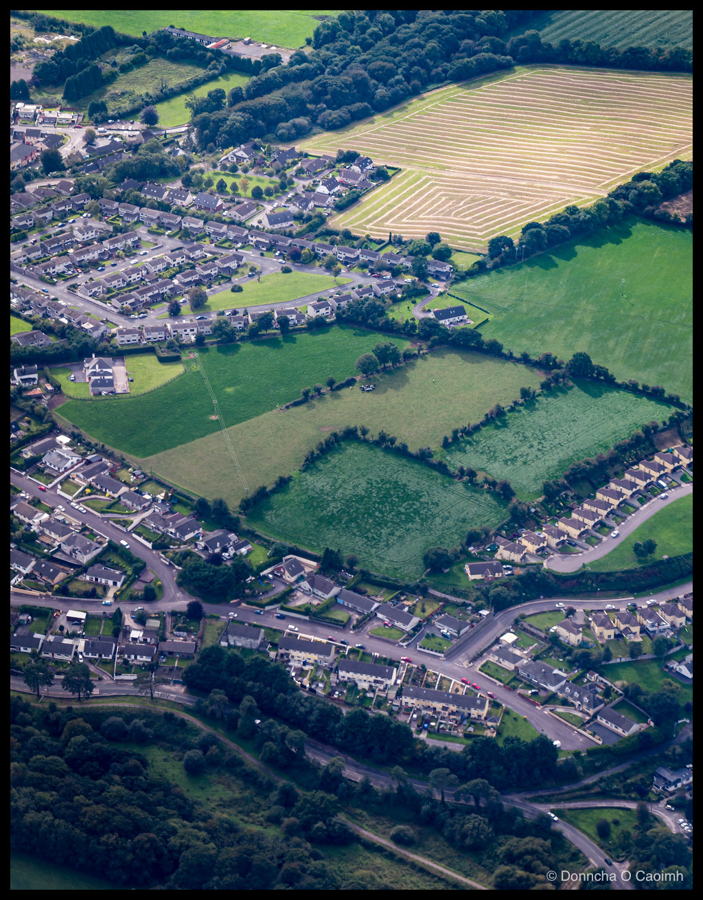 Aerial photo of a Blarney suburb in 2016, showing rows of detached houses bordering a large striped harvested field, with green pastures, hedgerows and woodland framing the scene.