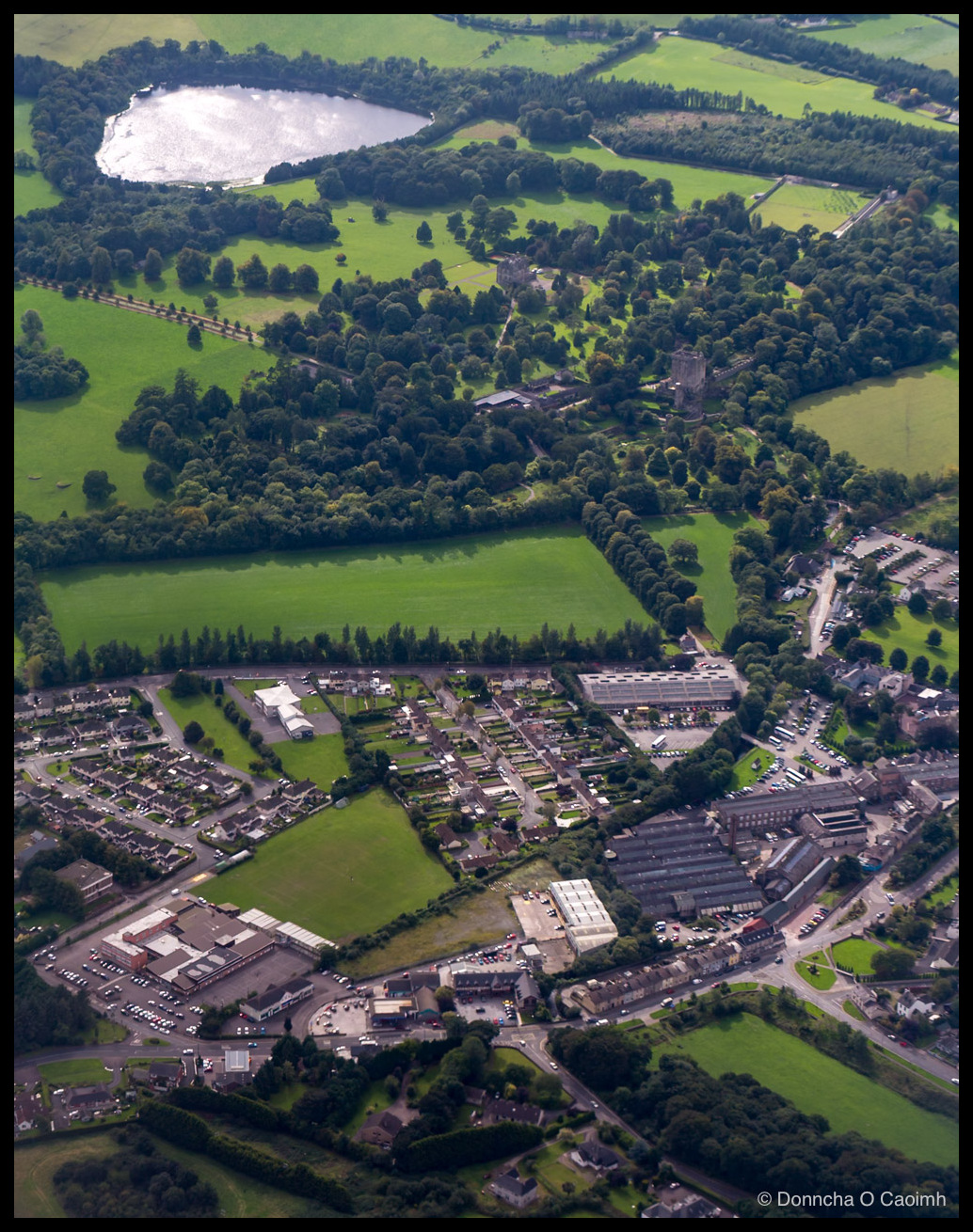 Aerial photo of Blarney village in 2016, with the secondary school and car park at lower left, a filling station and terraced housing in the middle, the Woollen Mills complex at right, and Blarney Castle, its grounds and Blarney Lough visible at the top.
