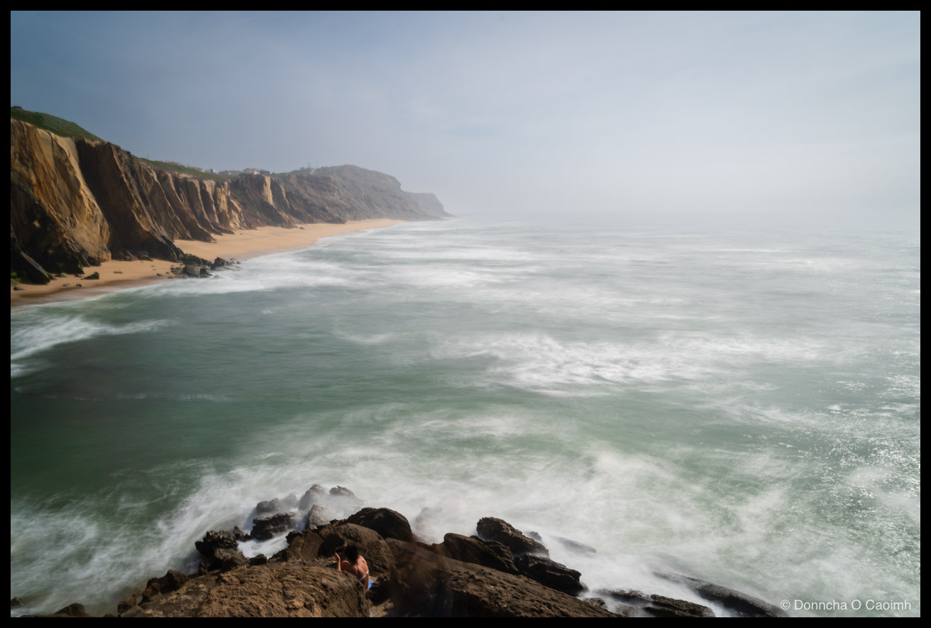 Long-exposure photo of a hazy coastline at Santa Cruz, Portugal, with tall ochre cliffs sweeping down the left edge into a pale sandy beach, while milky waves smear across the green Atlantic and a small figure sits on a blue towel on dark rocks in the foreground