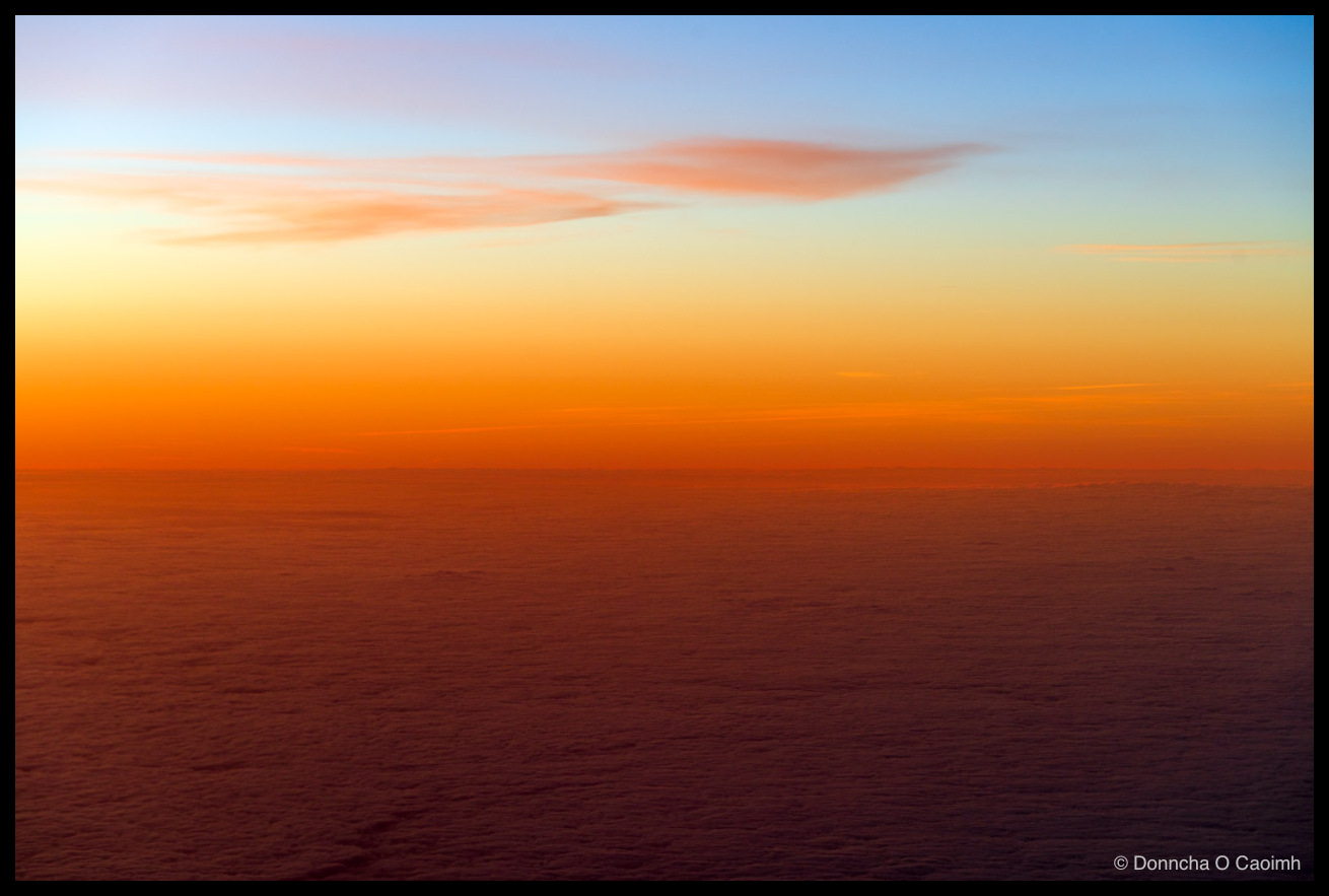 Sunrise viewed from an aircraft window, showing a wispy pink and grey cloud streak across a gradient sky that fades from soft blue at the top through orange to deep red near the horizon, above a vast textured carpet of cloud cover below.