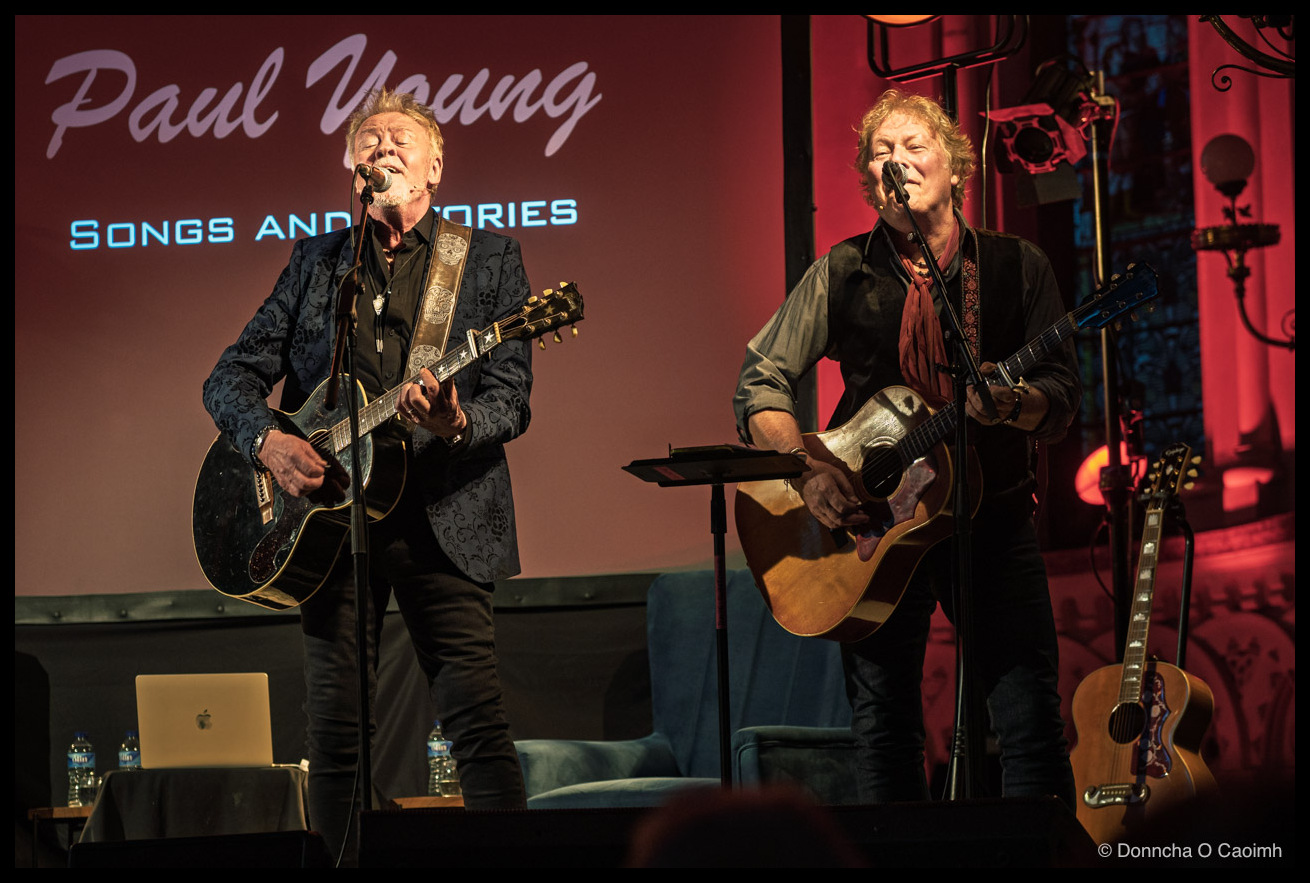 Two musicians performing on stage in front of a deep red projected backdrop reading “Paul Young” in cursive lettering with “Songs and Stories” beneath; the man on the left wears a blue paisley jacket and plays an electric guitar while singing into a microphone with eyes closed, the man on the right has grey curly hair, a dark shirt with a rust-coloured scarf, and plays an acoustic guitar; a vintage-style globe lamp glows top right, an acoustic guitar rests on a stand, and a MacBook and water bottles sit on a small table at stage left.