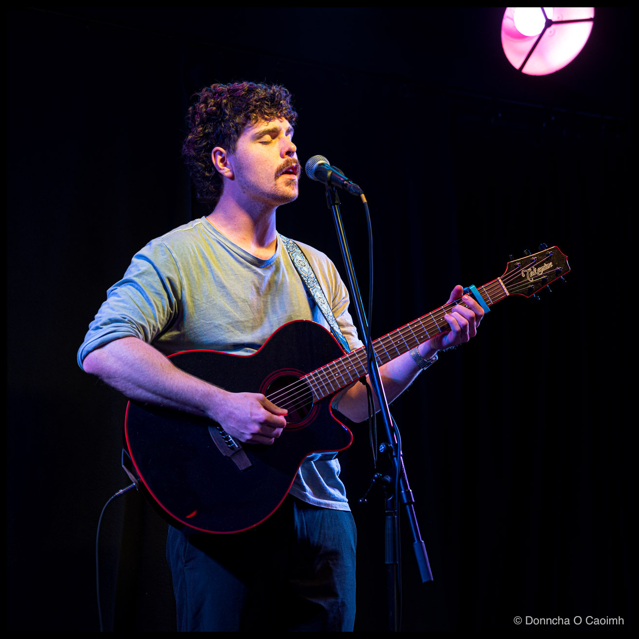 A curly-haired, moustached musician in a pale grey t-shirt, eyes closed as he sings into a microphone while playing a black Takamine acoustic guitar with red binding, a single pink stage light glowing in the upper right.