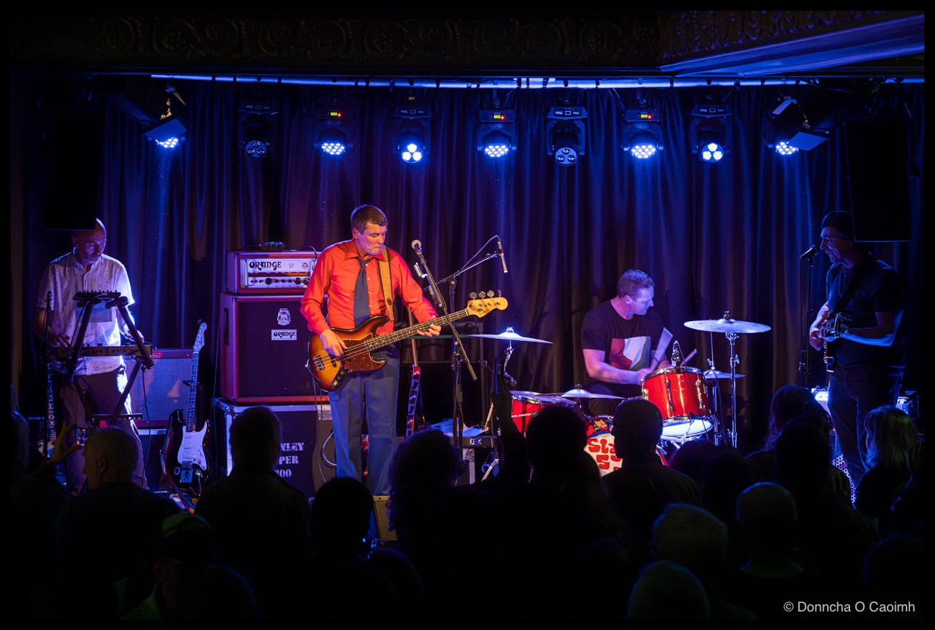 Photo of a four-piece band performing on a blue-lit stage above a crowd of silhouetted heads, with a keyboardist on the left, a bassist in an orange shirt and dark tie at centre, a drummer behind a red kit and a guitarist on
the right. Visible text: "ORANGE", "STANLEY SUPER 800".