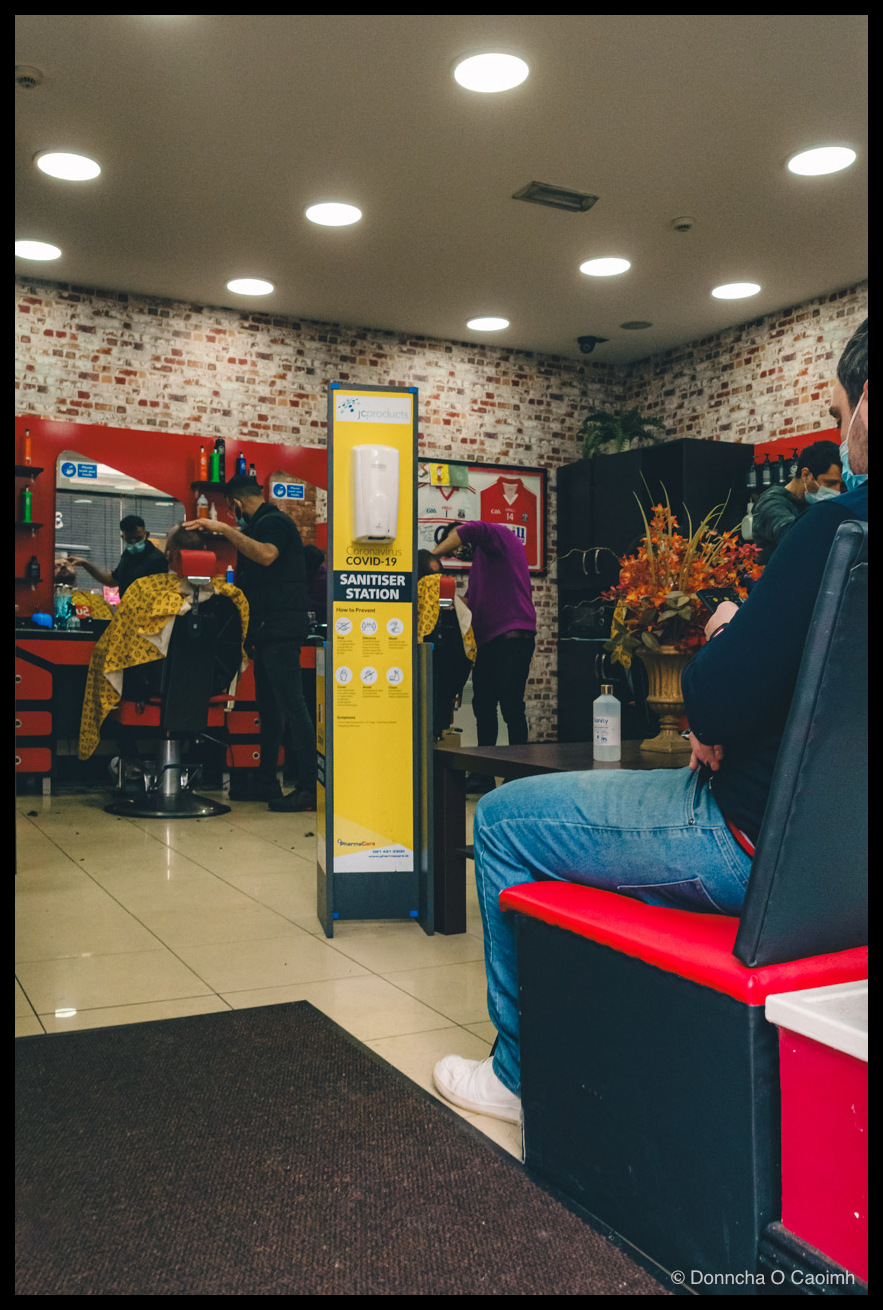 A Cork barber shop interior during the COVID-19 era: barbers in face masks cut hair for clients draped in yellow capes, with exposed-brick wallpaper, recessed ceiling lights and red-and-black furniture. A tall yellow "jcproducts Coronavirus COVID-19 SANITISER STATION How to Prevent" stand holds a hand-sanitiser dispenser in the centre, red Cork GAA jerseys (one numbered 14) hang on the wall, and a man in jeans and white trainers sits on a red bench in the foreground.