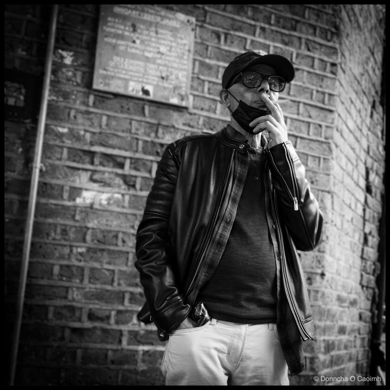 Black and white photo of a man leaning against a brick wall, smoking a cigarette with his face mask pulled down to his chin and one finger resting near his lips. He wears a flat cap, dark-rimmed glasses, an open leather jacket over a dark top, and pale trousers, with one hand in his pocket. A weathered metal plaque is mounted on the wall above his shoulder, its text too faded to read.